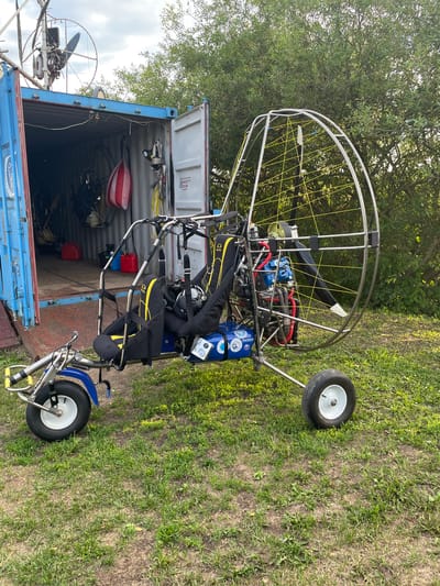 A powered paraglider cart sits on grass outside an open blue shipping container in Турово, Russia. The cart has two seats with yellow and black padding, a metal frame, and a propeller. Inside the shipping container, other equipment, including what appears to be another paraglider wing and red fuel cans, are visible. The container is blue with the doors open to the right, revealing the interior. Trees and foliage form the background, with a cloudy sky overhead. The scene is well-lit by daylight.