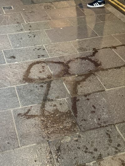 The image depicts a stone-paved surface with what appears to be a dark, dried liquid stain shaped roughly like a figure-eight or two overlapping circles with additional splatters and trails. A shoe with white stripes is visible in the upper left corner of the frame. The location is in Windermere, United Kingdom, evidenced by the street paving and street markings.