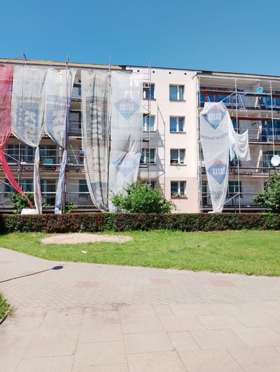 A light-colored, multi-story apartment building, covered in protective tarps and scaffolding, is visible under a clear blue sky. The building has several rows of windows and balconies. The tarps are white, with some featuring logos and text, including "ATLAS" and "MOŻESZ WIĘCEJ". A red tarp is also partially visible on the left side of the building. In front of the building, there is a neatly trimmed hedge, followed by a grassy lawn with a small mound of sand. A paved walkway, made of interlocking bricks, runs in the foreground. The overall scene suggests ongoing construction or renovation work on the apartment building. This image was taken in Kolno, Poland.
