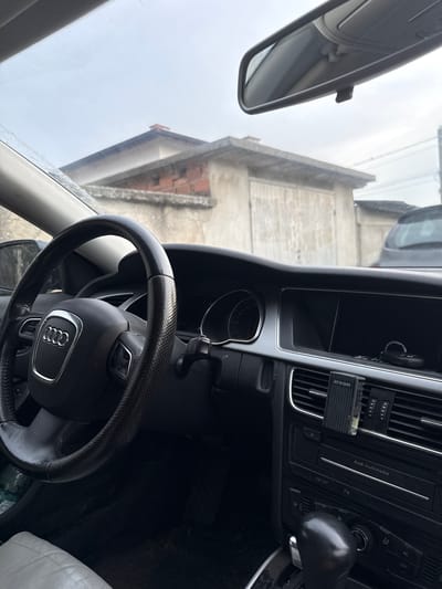 The image shows the interior of a car, likely an Audi, from the perspective of the driver. The steering wheel, with the Audi logo visible, is in the foreground. The dashboard features air vents, buttons labeled "ESP OFF," "P," and "AC OFF," and a multimedia system. An "areon" brand air freshener is clipped to an air vent. The rearview mirror is visible, reflecting an overcast sky and a building with a tiled roof, suggesting a cloudy day. The overall setting appears to be a parking area in Skutare, Bulgaria, given the location context provided. There is no clear indication of any activity or event taking place, other than potentially being parked. The mood is neutral, with no strong emotional cues conveyed. The image is taken from inside the car looking outwards and towards the dashboard.