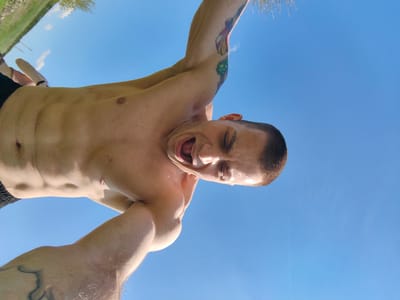 A shirtless man is angled upwards against a clear blue sky, with a hint of green grass and trees visible at the top left corner. He is fair-skinned, with a shaved head and tattoos on both arms. His mouth is open wide, and he appears to be yelling or screaming. He is likely in Neofit Rilski, Bulgaria, based on the user prompt.