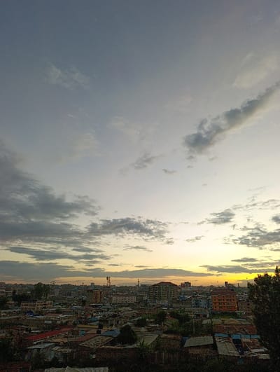 A cityscape under a cloudy sky with a visible sunset glow. Buildings of various heights are nestled closely together, reflecting the warm light of the setting sun. Dark shadows are cast in the foreground, suggesting a low vantage point overlooking the urban sprawl. The sky is filled with a mix of gray and white clouds, hinting at a recent rain or an approaching storm.
