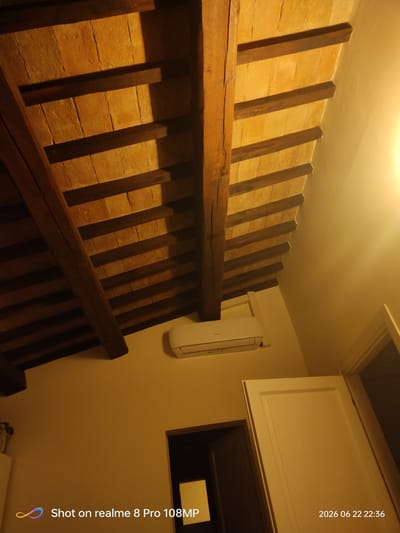 The image depicts the interior of a room with a traditional Italian architectural style, characterized by exposed wooden beams and terracotta tiles forming the ceiling. A white air conditioning unit is mounted on the wall beneath the ceiling. In the lower part of the image, a partially open dark wooden door is visible, and to its right, a portion of a closed white door is present. The overall lighting suggests an indoor environment with artificial light sources. The image is overlaid with text in the bottom left corner that reads "Shot on realme 8 Pro 108MP" and in the bottom right corner, a date and time stamp "2026 06 22 22:36". Without additional context or specific visual cues within the image that tie it to Foligno, Italy, its precise location cannot be confirmed from the image alone.