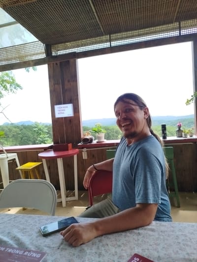 A man with long brown hair tied back and a beard is smiling and looking at the camera while sitting at a table on a covered patio. He is wearing a blue t-shirt and olive-colored pants. On the table in front of him is a smartphone and a placemat with floral patterns. Behind him, a red table and a yellow stool are visible. The patio overlooks a lush green landscape with mountains in the distance. A sign on a wooden post reads "TIỆM KEM TRONG RỪNG," which translates to "Ice cream shop in the forest." The scene appears to be in Trường An, Vietnam.