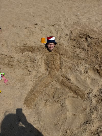 A person is buried up to their neck in sand on a beach. They are wearing a bucket hat with red, white, and blue stripes. The person's eyes are closed and they appear to be smiling. The sand is wet in some areas and dry in others. There are several children's beach toys scattered around the sand, including a yellow bucket and a pink shovel. The shadow of a person holding a phone is visible in the bottom left corner of the image, suggesting they are taking the photo. The scene appears to be taking place on a sunny day at the beach in Vityazevo, Russia.