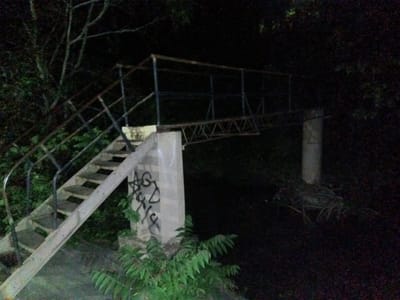 The image depicts a deteriorated bridge at night in Tahlequah, Oklahoma, United States of America. The bridge structure features a metal railing and a wooden staircase leading to the elevated walkway. Concrete supports, marred with graffiti, uphold the bridge. Vegetation is visible around the base of the bridge supports, while dense foliage surrounds the scene, obscuring the background in darkness. There are no people visible in the image.