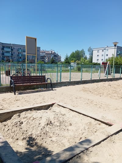 The image depicts an outdoor public area, likely a playground, in the city of Radzymin, Poland. In the foreground, a large sandbox filled with light brown sand occupies a significant portion of the frame. The sandbox is bordered by wooden planks, with one plank appearing to be a more recent addition or repair, marked with pink tape. Beyond the sandbox, a dark brown wooden bench with black metal legs sits on the sand. Behind the bench, a green metal fence encloses a paved area with some cylindrical bollards and bicycles. Further back, a large, weathered wooden sign with a grey surface stands in front of residential buildings. The background consists of several multi-story apartment buildings, some with balconies, and a clear blue sky. Trees and a street lamp are also visible. The overall scene suggests a sunny day in a residential urban environment.