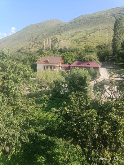 The image depicts a rural landscape in Rakhata, Russia, on a sunny day. In the foreground, lush green trees and foliage dominate the scene, casting dappled shadows. Behind the trees, two buildings with red tiled roofs are visible. The larger building on the left is a multi-story house with a light-colored facade and balconies. The smaller building to its right appears to be a single-story structure. A narrow bridge spans a dry riverbed or shallow stream in front of the buildings, with a dirt road leading away from them. In the background, a large, gently sloping mountain covered in green vegetation rises under a clear blue sky with a few scattered clouds. Several tall, thin trees are scattered across the mountainside. A white vehicle is parked on the side of the road near the smaller building. The timestamp "10.07.2026 16:24" is visible in the lower right corner of the image.