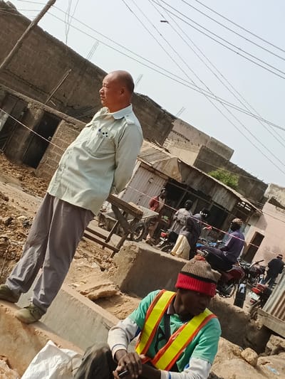 The image depicts an outdoor scene in Gobirawa, Nigeria, under a bright, clear sky. In the foreground, positioned to the left, an East Asian male with a bald head stands facing right, his hands clasped behind his back. He wears a light khaki-green collared shirt with two breast pockets, one containing pens, grey trousers, and olive-green shoes. To the lower right, an African male sits on a concrete block, wearing a red and black knitted beanie hat, a light green long-sleeved shirt, and a yellow reflective safety vest. He is looking down, holding a dark object, possibly a mobile phone.

The background features multiple rudimentary buildings constructed from concrete blocks, some appearing unfinished or partially dilapidated, with exposed rebar in one structure on the far left. Corrugated metal roofing is visible on some smaller structures. The ground is unpaved, uneven, and appears dusty with scattered debris. Several other individuals, all appearing to be of African descent, are present in the mid-ground. One person stands near a wooden handcart, while another sits on a red motorcycle. Multiple motorcycles are parked or being operated in the distance. Overhead wires, likely electrical or communication cables, crisscross the upper portion of the frame.