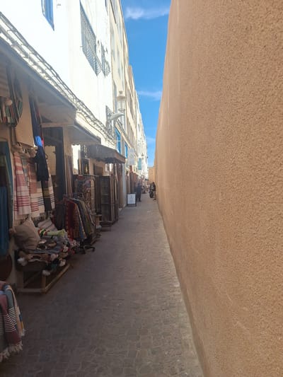 This image depicts a narrow street in Essaouira, Morocco, lined with shops on the left and a textured, light brown wall on the right. The street is paved with cobblestones. On the left, several market stalls display textiles, including striped scarves and patterned fabrics, along with woven baskets and decorative items. A few people are visible walking down the street in the distance. The buildings on the left have white facades with blue-shuttered windows and wrought-iron bars. The sky above is a clear blue with a few wispy white clouds.