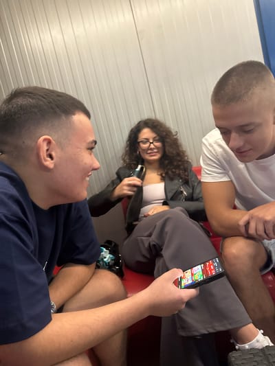 Three individuals, two males and one female, are seated on a red couch. The male on the left is wearing a dark blue t-shirt and a watch, looking to his right with his mouth open in a smile. The male on the right is wearing a white t-shirt and looking down at his hands. The female in the middle is wearing a black leather jacket over a light-colored top and dark pants, holding a vape device. She is wearing glasses and looking forward. In front of the male on the left, a smartphone is visible, displaying colorful app icons. The background consists of ribbed, light-colored walls. The location is identified as Okop, Bulgaria.