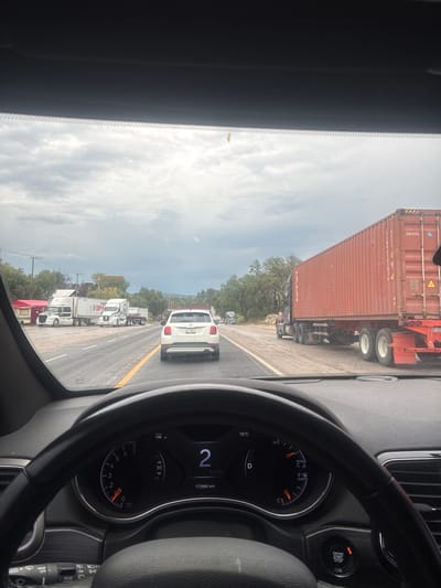 The image shows a view from the driver's seat of a vehicle, looking forward through the windshield. The vehicle is on a wet asphalt road, suggesting recent rain. Traffic is present, including several semi-trailer trucks, one of which is a large red shipping container truck. A white SUV is directly ahead in the lane. The dashboard of the vehicle is visible, showing a speedometer reading 2 km/h and an odometer reading 173990 km. The sky is overcast with heavy clouds. The surrounding environment appears to be rural or semi-rural, with trees and some roadside structures, including a red food stand. The context of Tepeji de Ocampo, Mexico, is provided, though not visually evident within the image itself.