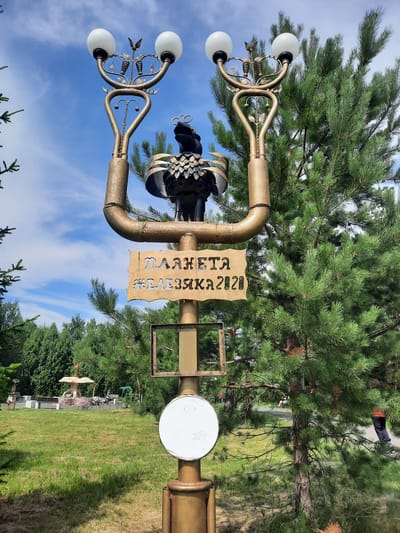 A metal sculpture resembling a candlestick with a bird-like figure at the top is mounted on a pole. Below it, a wooden sign with cut-out Cyrillic letters reads "ПЛАНЕТА ЖЕЛЕЗЯК 2020" (Planet of Iron 2020). Two empty square frames are attached to the pole below the sign, and a circular white sign is mounted further down. The sculpture is set in a park-like environment with lush green grass, mature pine trees, and other metal sculptures in the background, including a zebra. The sky is blue with scattered clouds. The location is identified as Zelenogorsky, Russia. There are a few people visible in the background, walking on a path and sitting on benches. The overall scene suggests an outdoor art installation in a public park.