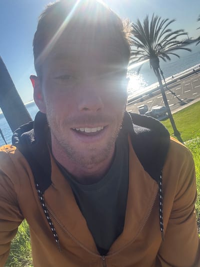 A sun-drenched selfie shows a man with short blond hair and a light beard, smiling. He is wearing a brown jacket over a green shirt. Behind him, a palm tree and a bright blue ocean are visible. The sun reflects off the water, creating a sparkling effect. A road and green grass run alongside the water. The image is rotated.