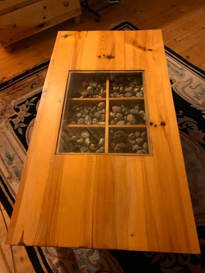 In a dimly lit room, a wooden coffee table with a glass-covered inset displaying an assortment of seashells and small stones is centrally positioned on a patterned rug. The table, made of light-colored wood, has a rustic appearance with visible grain and knots. The inset is divided into six compartments by wooden dividers, each filled with shells and stones of varying shapes and colors. The rug beneath the table features a dark background with intricate floral and swirling patterns in lighter shades. In the background, a wooden dresser with drawers and wooden flooring are visible. The scene is devoid of any people. The location is Mepiskalaki, Georgia.