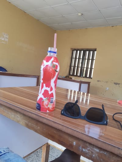 The image depicts an indoor scene, likely a classroom or common area, in Palade, Nigeria. A wooden table occupies the foreground, upon which rests a bottle of strawberry-flavored juice with a pink straw. Next to the bottle, a pair of black sunglasses is placed. The wall behind the table is painted a pale yellow and shows some signs of wear and tear, including peeling paint and what appears to be graffiti or writing. A barred window is visible on the wall, suggesting the building's security features. The ceiling is made of white tiles. A glimpse of a person's blue jeans can be seen in the lower left corner of the frame, indicating their presence.