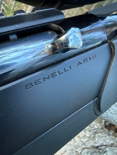 The image shows a close-up of a black shotgun with the inscription "BENELLI ARMI" on its side. The shotgun is resting on a wooden surface. The inscription is clearly visible and embossed into the metal. The lighting suggests it is daytime. There is no indication of any city or location within the image itself.