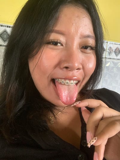 In a close-up selfie, a young Indonesian woman with black hair and braces playfully sticks out her tongue. Her eyes are dark and her lashes are long and accentuated. She wears a black top and a delicate gold necklace. Her fingernails are painted a pale pink with black polka dots. The background shows a yellow wall and patterned tiles. The image appears to have been taken indoors, possibly in a home environment in Jajag, Indonesia, given the distinctive yellow wall and tiled background often seen in residential settings in the region.
