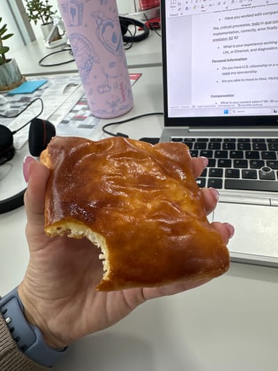 An overhead view captures a person's hand holding a golden-brown, glazed pastry with a bite taken out, set against a cluttered desk in what appears to be an office or home office environment. The hand, belonging to a woman, has manicured nails with a light pink or clear polish and wears a light blue smart watch with a silicone strap on the left wrist.

The primary activity is the person holding and presumably eating the pastry, indicating a break or snack while working. The pastry has a glossy, somewhat textured glaze, and the exposed interior reveals a lighter, bready texture.

In the background, a silver laptop computer is open, displaying a document or online form on its screen. Visible text on the laptop includes questions and sections related to employment, such as "Have you worked with compan," "critical processes," "What is your experience working," "Personal Information," "Do you have U.S. citizenship or a va," "need any sponsorship," "Are you able to move to Novi, Michig," and "Compensation," with a specific query "What is your current salary? 80 - 90 nc." Other small text like "Paste," "Clipboard," "Font," "Paragraph," and a "Search" icon are also discernible.

Various items are scattered on the white desk surface. A pink mouse pad with intricate light blue patterns (hearts, cherries, bottles) and the "owala" logo is visible to the left of the laptop. Black headphones with red accents are also present, along with small potted plants, contributing to a busy workspace aesthetic.

The setting is an indoor location, with bright, even lighting, suggesting daytime. The contextual location of the image is Sanctorum, Mexico.