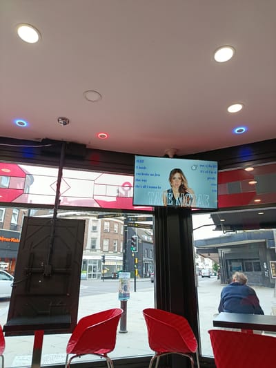The image depicts the interior of a business, likely a cafe or fast-food establishment, with a view of a street scene through large windows. Inside, there are several red chairs and a black stand holding a large screen. The screen displays a music video or lyrics, featuring a portrait of Tate McRae, along with song titles like "2 hands," "you broke me first," and "greedy." Overhead, recessed ceiling lights and colored LED lights (blue and red) are visible.
Through the windows, the exterior scene shows a typical urban street in Bedford, United Kingdom. Buildings of various architectural styles line the street, including shops with signage like "Döner Keb" and "Parity Outlet Shop." Traffic lights and street signs indicating directions to "The Higgins," "Allhallows," and "Harpur Centre" provide specific location cues. The weather appears to be overcast, with diffused natural light. A person is seen sitting at a table in the background, facing away from the camera, while a white van drives by on the street. The overall mood is ordinary, capturing a moment in a public space with elements of entertainment and daily life.