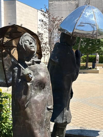This is a medium shot of the “Meeting” statue, a bronze sculpture by Hungarian sculptor Imre Varga in Siófok, Hungary. The statue consists of two older male figures dressed in coats, hats, and holding umbrellas. One figure has a cigar in his hand. The sculpture is set outdoors on a paved surface with buildings and trees visible in the background. The sky is clear.
