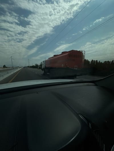 The image captures a view from inside a vehicle traveling on a highway. The highway has multiple lanes and is bordered by a concrete barrier on the left. In the distance, several vehicles are visible on the highway. To the right of the highway, there are trees and power lines supported by tall pylons.
Dominating the right side of the frame is a large, red tanker truck. The truck has a cylindrical tank with some writing and a symbol on its side. The sky is a mixture of blue with scattered white clouds, and several dark, thin power lines run diagonally across the upper portion of the image. The foreground shows the dark dashboard of the vehicle from which the photograph is taken. The scene appears to be in a semi-urban or rural area, given the presence of the highway, pylons, and vegetation. The overall impression is one of travel on a clear, albeit partly cloudy, day.
The location is the city of Istiqlol, Uzbekistan.
