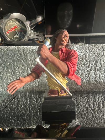 The image shows a close-up view of a detailed figurine of a man singing into a microphone. The figurine is wearing a red open shirt over a yellow garment and a brown belt. He has long dark hair and his mouth is open in a singing expression. The figurine is placed on a black base. In the background, there is a motorcycle-shaped clock, with the brand "CBR" visible. Further back, a blurred reflection of a face is visible on a dark surface. The surface on which the figurine and clock are placed appears to be textured and grey. The setting is indoors, with lighting that creates highlights and shadows. There is no indication of the city of Brca, Montenegro, in this image.