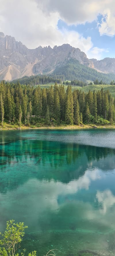 This image captures a scenic view of the Karersee (Lago di Carezza) in Welschnofen - Nova Levante, Italy. The foreground is dominated by the crystal-clear, emerald-green waters of the lake, reflecting the sky and surrounding landscape. The clarity of the water allows visibility of the lakebed with submerged elements like logs and aquatic vegetation. On the far side of the lake, a dense forest of tall evergreen trees lines the shore, transitioning into a backdrop of imposing, rugged mountains. The mountains exhibit a rocky, textured appearance with some patches of snow or lighter-colored scree, and their peaks are partially obscured by clouds. The sky is a mix of bright clouds and patches of blue. The overall impression is one of natural beauty and tranquility in a mountainous alpine setting. There are no people or specific actions depicted in the image; it is a landscape photograph.