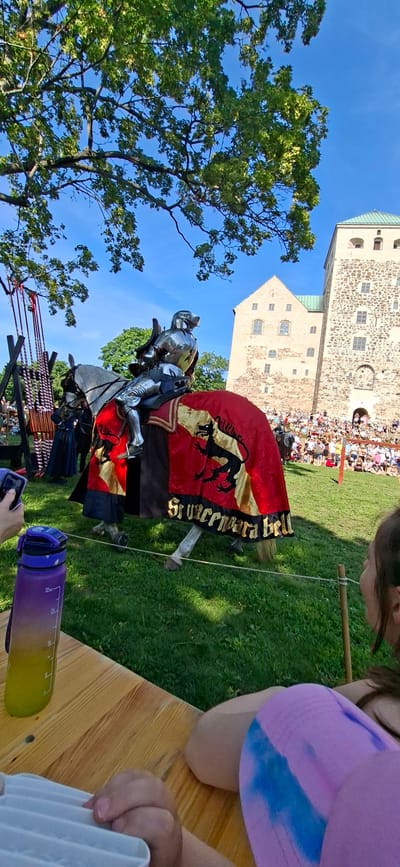 The image shows a reenactment of a jousting tournament at Turku Castle in Turku, Finland. A knight in full armor is mounted on a horse, which is adorned with a red and gold cape featuring a black dragon emblem and the inscription "Si Vincemara Bell". In the background, a crowd of spectators watches the event, with the stone walls of Turku Castle visible behind them. The scene takes place on a grassy field under a clear blue sky, with a large tree casting a shadow over the area. In the foreground, a wooden table holds a purple water bottle and a white object, likely a phone or a food container. A person's hand is visible holding the white object, and another person's face is in the lower right corner of the frame.