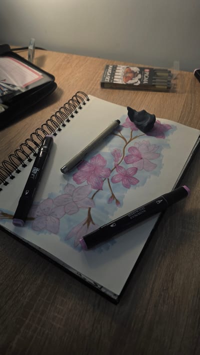 The image shows a close-up, eye-level view of a wooden desk with art supplies. A spiral-bound sketchbook lies open, displaying a drawing of pink cherry blossoms with light blue shading. Three markers are placed on the sketchbook: a black marker with purple accents on the left, a gray marker in the center, and another black marker with purple accents on the right, partially obscuring the drawing. A small piece of dark gray putty or kneaded eraser rests on the drawing, near the center. In the background, to the left, is a black zippered art supply case, partially open, revealing pink and white paper inserts. To the right of the sketchbook, a small cardboard box containing Pigma Manga markers is visible, with the label "MANGA SET" clearly legible. The lighting suggests an indoor setting, possibly a desk lamp illuminating the scene, casting shadows on the wooden surface. There are no people visible in the image. The scene is static and appears to be a workspace for drawing or sketching. The location is not discernible from the image.