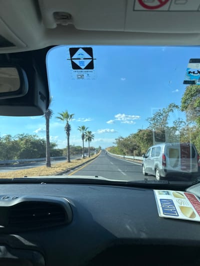 View from inside a car driving on a sunny road. Palm trees line the roadside. A white van is visible ahead. Stickers are on the windshield. The car's interior features a dark dashboard and light-colored trim. Clear blue sky with scattered clouds is seen through the windshield.