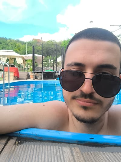 In an outdoor setting, a man is in a swimming pool. He is wearing aviator sunglasses and has a short haircut and a beard. The pool is filled with blue water, and the edge of the pool is visible, appearing to be made of blue plastic or painted wood. In the background, there are trees, a trampoline, gazebos, and what appears to be a building. The sky is blue with white clouds. The location is Stob, Bulgaria, likely a private residence or a recreational area given the presence of a pool and outdoor amenities.
