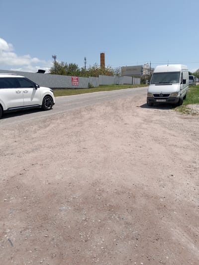 The image depicts a rural road in Rayevskaya, Russia. A white Kia SUV is parked on the left side of the road, while a white Mercedes-Benz van is parked on the right. A corrugated metal fence runs along the roadside, with a sign posted on it. In the background, there is a brick chimney, cell towers, and a building labeled "ROYAL HALL". The road is unpaved and covered in dirt and gravel. The sky is clear and blue.