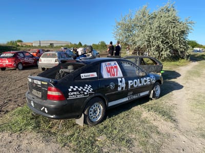 This image depicts a dirt track race in the Kanjiza Municipality, Serbia. Prominently featured is a black Opel Vectra race car marked with "54 Police" and the number 407, parked on a dirt track next to a tree. The car has racing modifications like a roll cage and a metal mesh covering the rear window. Other race cars are visible in the background, including red and white cars with racing decals and numbers. Several people are standing near the cars, one group appears to be wearing helmets. The background includes a building and some trees. The overall environment suggests a rural or semi-rural setting where the dirt track race is taking place.