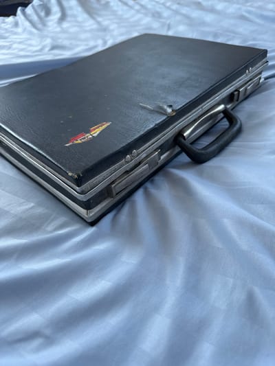 A black briefcase rests on a blue, subtly striped fabric surface, possibly a bedspread or tablecloth. The briefcase is closed and angled, with its silver-toned trim visible along the edges. A black, plastic handle is attached to the right side of the briefcase. On the surface of the briefcase, there is a small, colorful sticker depicting a red, yellow, and white stylized object, possibly a boat or a flag. There is also a small tear on the top surface of the briefcase. The lighting creates subtle shadows and highlights on the fabric. The image does not contain any elements that would suggest a location within the city of Urazovo, Russia.