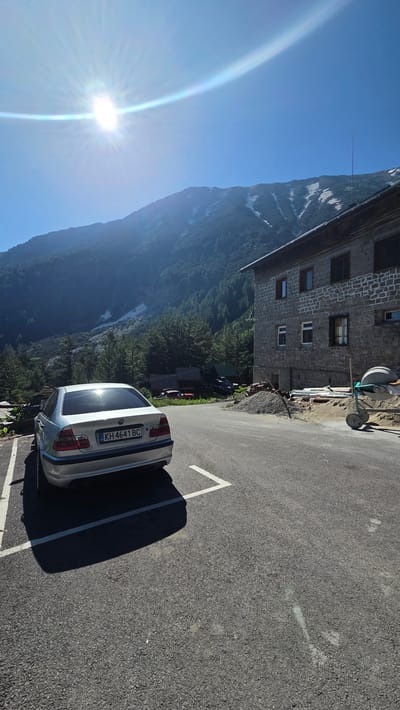 A silver BMW sedan is parked in a lot on a sunny day. The car is facing away from the camera, with its rear license plate, KH 4641 BC, visible. The parking lot is paved and has white parking lines. In the background, a stone building with multiple windows stands next to a pile of gravel and construction materials, including a cement mixer. Beyond the building, a lush green forest climbs up a mountainside. The mountain peak is capped with patches of snow. The sky is a clear, vibrant blue with a bright sun high in the sky, casting a lens flare and a rainbow-like prism effect.