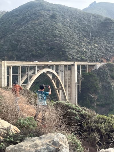 Bixby Bridge dominates the scene, a concrete structure with prominent arches and supports. It spans a gap between a steep, vegetated hillside on the left and a rocky, shrub-covered area on the right. Two figures are visible. One in a blue shirt is poised as if taking a photograph. A second figure, in an orange shirt, lies prone further down the hillside, also seemingly taking a photo. The sky is overcast, contributing to a muted, naturalistic color palette.