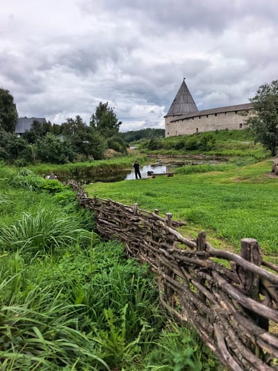 A person stands near a body of water in Staraya Ladoga, Russia, beside a rustic wooden fence. The scene depicts a historical fortification, likely the Staraya Ladoga Fortress, characterized by its stone walls and a conical wooden tower. Lush green grass covers the landscape, with trees and foliage surrounding the water and buildings. The sky is overcast with heavy clouds, suggesting a potentially cool or rainy day. The foreground is dominated by the textured, weathered wood of the fence, while the background features the imposing fortress architecture.