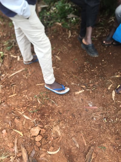 This image depicts an outdoor ground scene in Rigoma ward, Kenya, focusing on the lower extremities of three individuals standing on reddish-brown soil.
The ground is unpaved, consisting primarily of dry, cracked reddish-brown soil. Scattered across the surface are various natural debris elements, including numerous dried leaves, some light in color and others dark, along with small twigs and fragments of wood. Several small, irregular-shaped rocks are also visible on the ground. Sparse, small green plant sprouts are present in various locations, indicating minimal vegetation. A small patch of white, powdery substance is discernible on the ground towards the middle-right.
From left to right, the visible lower body parts of three individuals are present. The leftmost individual is wearing light-colored (possibly khaki or cream) pants. Their right foot is visible, clad in blue and white flip-flops with a blue strap and a white and brown sole. To the right, another individual wears black pants and black closed-toe clogs or slip-on shoes. Further to the right, only the lower leg and foot of a third person are partially visible, wearing dark-colored (likely black) flip-flops. All individuals appear to be standing still, with no active interactions or specific actions discernible beyond their presence on the scene. The background beyond the ground level is out of focus but appears to be dense green foliage.