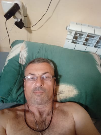 The image shows a man lying in bed, propped up against a green pillow with a feather design. He is wearing glasses and a necklace. To the left of the bed, a wall outlet is overloaded with multiple plugs and adapters. To the right, a white radiator is visible. The overall environment suggests an indoor setting, likely a bedroom, in Saky, Ukraine.