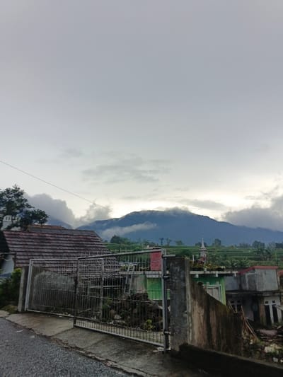 The image depicts a rural mountainous landscape in Sindangjaya, Indonesia, under an overcast sky. The foreground features a dark asphalt road or path on the left, alongside a concrete curb and a partially open metal gate with concrete posts. Behind the gate, a structure with a reddish-brown tiled roof is visible. To the right, a concrete wall stands. In the midground, several houses with varied roof colors (green, grey) and construction styles are observed, some appearing incomplete or in disrepair. Beyond these residences, terraced agricultural fields, likely rice paddies, ascend the lower slopes. A small, white-spired structure resembling a miniature mosque minaret is present among the houses and fields on the right. The background is dominated by large mountains, their peaks and upper slopes partially shrouded in dense, low-hanging clouds and mist. The sky is a uniform light grey, characteristic of overcast conditions, with a subtle brighter area on the right horizon partially obscured by clouds. A faint, dotted utility line horizontally crosses the upper portion of the frame. No individuals are visible, and no actions or interactions are depicted.