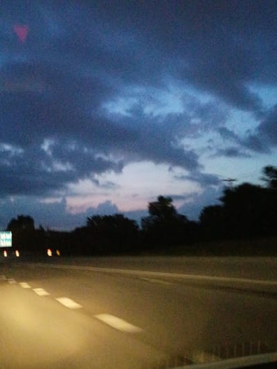 The image shows a blurry, low-light view of a highway at dusk or dawn. The sky is filled with dark, textured clouds, with lighter patches indicating the fading light of sunset or sunrise. Silhouetted trees line the horizon. The road in the foreground is asphalt, with visible white lane markings that are illuminated by the headlights of an unseen vehicle. To the left of the road, a traffic sign with green and white text is partially visible. The overall impression is one of a transitional time of day on a highway, possibly in a rural or suburban area adjacent to a city. The image does not provide enough detail to identify a specific location within Lexington, United States of America.