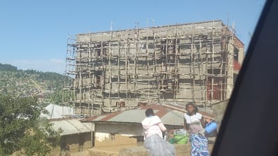 A tall building under construction is covered in scaffolding against a clear blue sky. Below, rooftops of smaller dwellings extend towards a hillside with scattered buildings. Two women stand near the structures; one sits wearing a light pink top and dark skirt, while the other stands in a white top and patterned blue skirt, holding a blue container. A vehicle's interior, partially visible, obscures the view on the right.