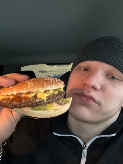 A male individual is positioned in the right foreground, partially obscuring the view of the background. He wears a dark grey knitted beanie and a black zip-up jacket with a white zipper, displaying a "NIKE" logo on the zipper pull. His blue eyes are visible, and he has light facial hair and a potential lip piercing on the lower right side of his mouth. He is holding a double cheeseburger in his left hand, presenting it towards the viewer. The burger consists of a sesame seed bun, two beef patties, melted yellow cheese, sliced pickles, and an orange/pinkish sauce. The bottom bun shows an irregular edge, possibly from a bite. The background suggests the interior of a vehicle, with a dark headliner and a rear window showing a dimly lit exterior with faint horizontal reflections, indicative of a night or dusk setting with potential snow or ice outside. The precise location within Div'ya, Russia, is not discernible from the image content.