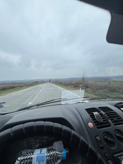 The image captures a view from inside a vehicle near Ovcha Mogila, Bulgaria, looking forward through a windshield onto a rural road under an overcast sky. The two-lane asphalt road, appearing damp and showing minor surface wear, recedes into the distance with a gentle curve to the left. White solid and dashed lines mark the road's edges and center. On the immediate right side of the road, green vegetation, a bare deciduous tree, and areas of disturbed earth with cut timber or brush are visible, along with red and white roadside markers. The landscape beyond features rolling hills and fields under a uniform grey sky.
Inside the vehicle, the dark dashboard occupies the lower portion of the frame. A black, textured steering wheel is partially visible on the left, concealing most of the instrument cluster, though a speedometer with markings up to 200 km/h is discernible. A clear plastic water bottle, labeled "5 LE" and fitted with a blue cap, rests horizontally on the dashboard in front of the steering wheel. To the right of the steering wheel, black air vents with a magnetic phone mount attached to one, and a control panel with various buttons and knobs, are present. A red circular symbol is also visible near the vents. The windshield shows minor reflections and surface residue. No individuals are visible within the frame.