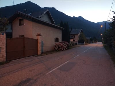 The image depicts a street scene in Saint-Étienne-de-Cuines, France, at dusk. A residential house with a brown gate and a stone pillar is visible on the left. In the background, a dark mountain range dominates the skyline under a pale sky. A road stretches into the distance, lined with streetlights and trees. The overall atmosphere is serene and quiet, suggesting the end of the day.