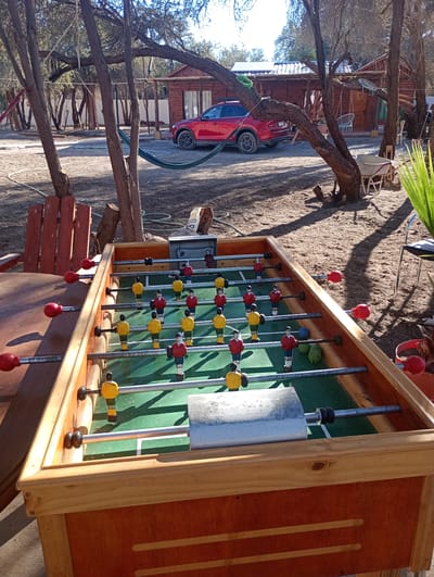 This image captures an outdoor scene centered around a wooden foosball table under bright daylight. The foosball table, occupying the foreground, features a green playing field with miniature red and yellow players arranged on metal rods. Red handles are visible on the left side of the table, ready for play.
The setting appears to be a recreational or residential area outdoors. Numerous trees with bare, sprawling branches are scattered throughout the midground and background, casting long shadows on the dry, sandy ground. A red SUV is parked in the midground, near a rustic wooden cabin or house with a porch. The cabin's roof appears to have a corrugated texture and possibly solar panels. A green hammock is strung between two trees, and a wooden chair with red slats is partially visible on the left. Further back, more trees and a white fence line the perimeter.
The scene is bathed in sunlight, suggesting a clear day, likely morning or afternoon given the shadow length. No people are visible, implying a quiet moment in this leisure space. The dry ground and sparse foliage are characteristic of an arid environment, consistent with the location context of Pozo Almonte, Chile. Other visible objects include a wheelbarrow and coiled hoses on the ground. No discernible text is visible on any objects in the image. The overall impression is one of an inviting, relaxed outdoor recreational area.
