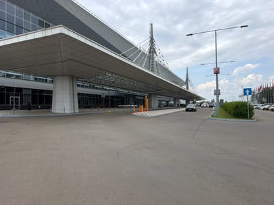 The image shows an exterior view of the airport terminal in Yemelyanovo, Russia. The modern architectural design of the terminal features a large, cantilevered roof supported by pillars and cables, with a glass facade. In the foreground, there is an asphalt road and pavement. Several cars are visible on the road and in a parking area to the right, where a row of flags can also be seen. A lamppost with a "P1" parking sign is present on the right side of the image. The sky is overcast with clouds. People are walking near the entrance of the terminal and by the cars. Traffic barriers are set up at the entrance.