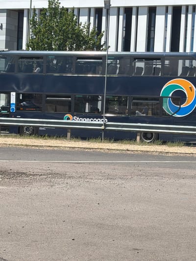 A dark blue double-decker bus with the Stagecoach logo is parked on a roadside in Tewkesbury, United Kingdom. The bus has tinted windows and several passengers are visible inside. The Stagecoach logo, a colorful swirl of orange, green, and blue, is prominently displayed on the side of the bus. In the background, a modern building with a striped facade and trees can be seen. The foreground shows a paved road with some gravel and debris scattered on it. A metal barrier runs along the side of the road.