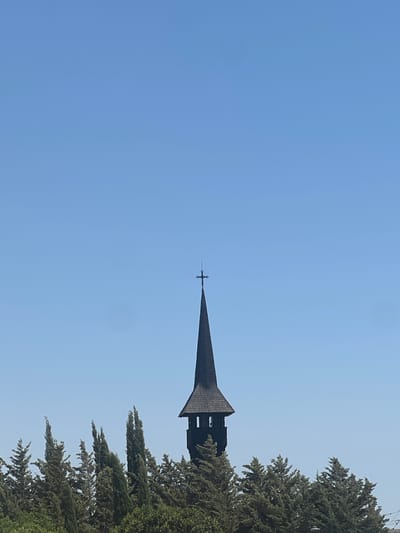 An image depicts a dark, pointed church steeple with a cross on top, silhouetted against a clear blue sky. The steeple is centrally located, rising above a dense line of green trees. The trees vary in shape and height, with some appearing tall and slender, possibly cypress trees, while others are more rounded and bushy. The overall impression is of a peaceful, natural setting with a prominent religious structure.